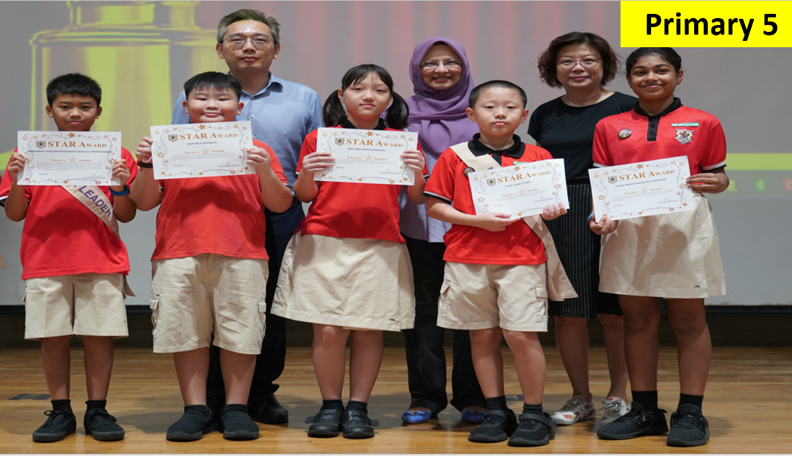 Five Primary 5 students in red uniforms hold Star Award certificates, flanked by two adult teachers on a stage.
